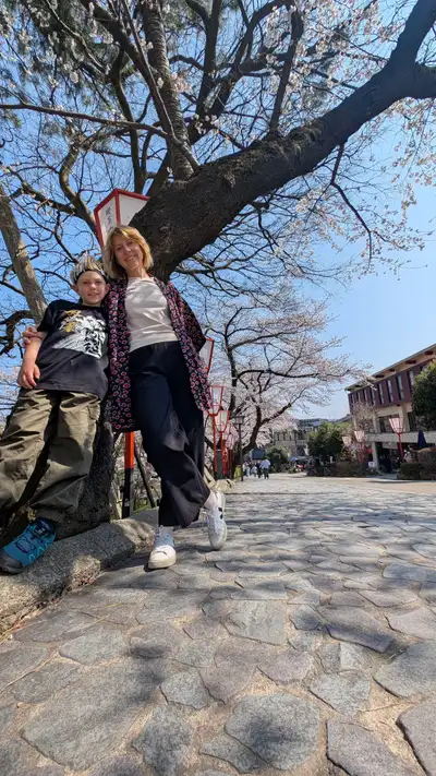 Dasha and Charlie leaning back against the trunk of an old cherry, shot from ground level looking up the stone-paved avenue. A line of red paper lanterns on poles runs off into the distance under trees in full blossom, clear blue sky. Dasha in a patterned haori over a white tee, Charlie in a black graphic tee and a white headband, cargo trousers, arms crossed.